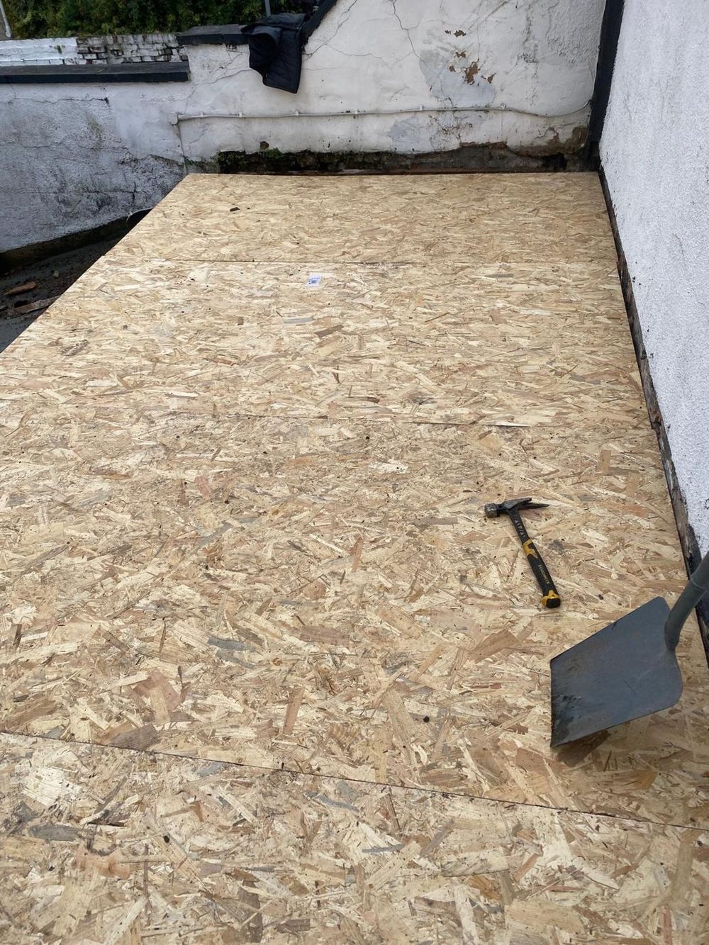 A new OSB deck laid out across the full width of a flat roof and run up to the rendered wall at the back, hammer and shovel still on it