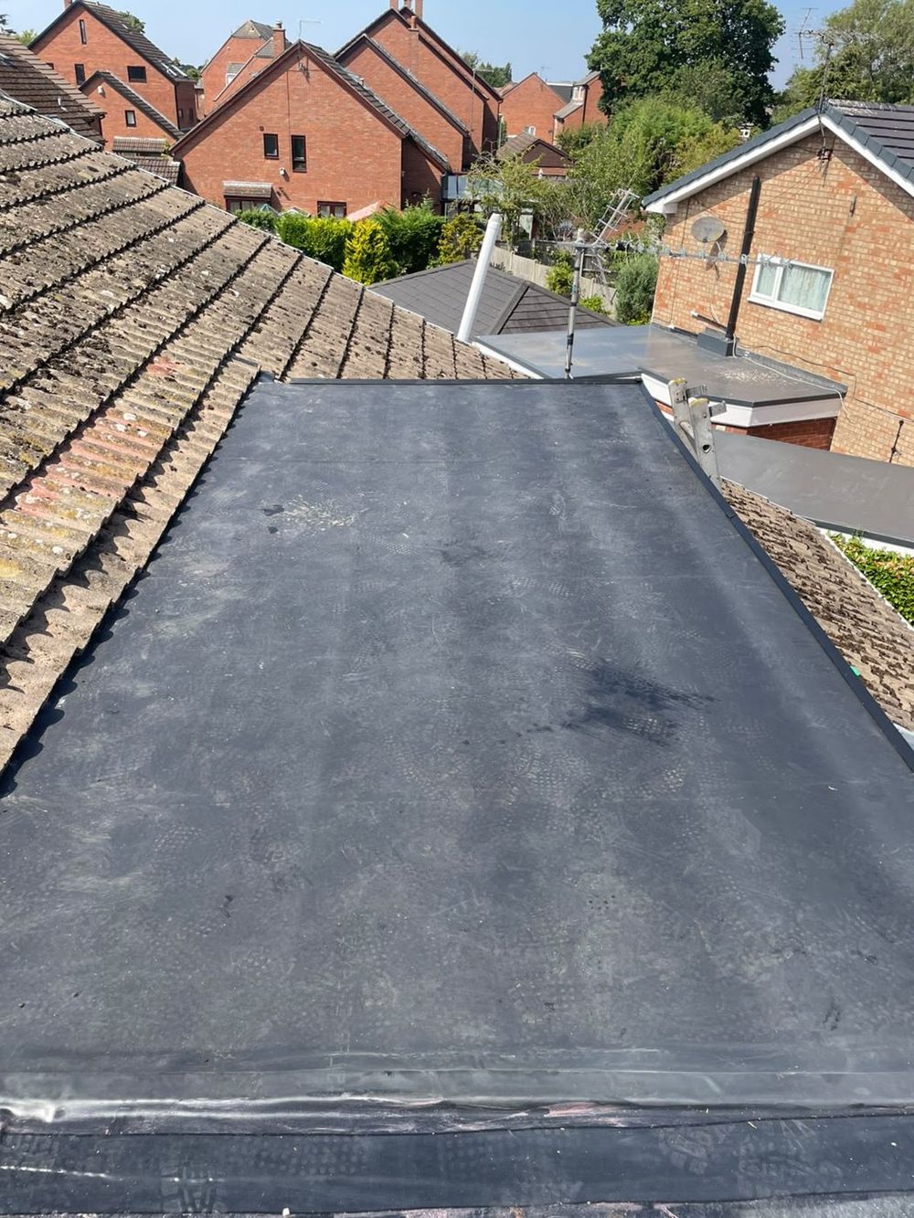 A finished rubber roof at upper level with a tiled slope rising on either side of it, seen from above the neighbouring rooftops