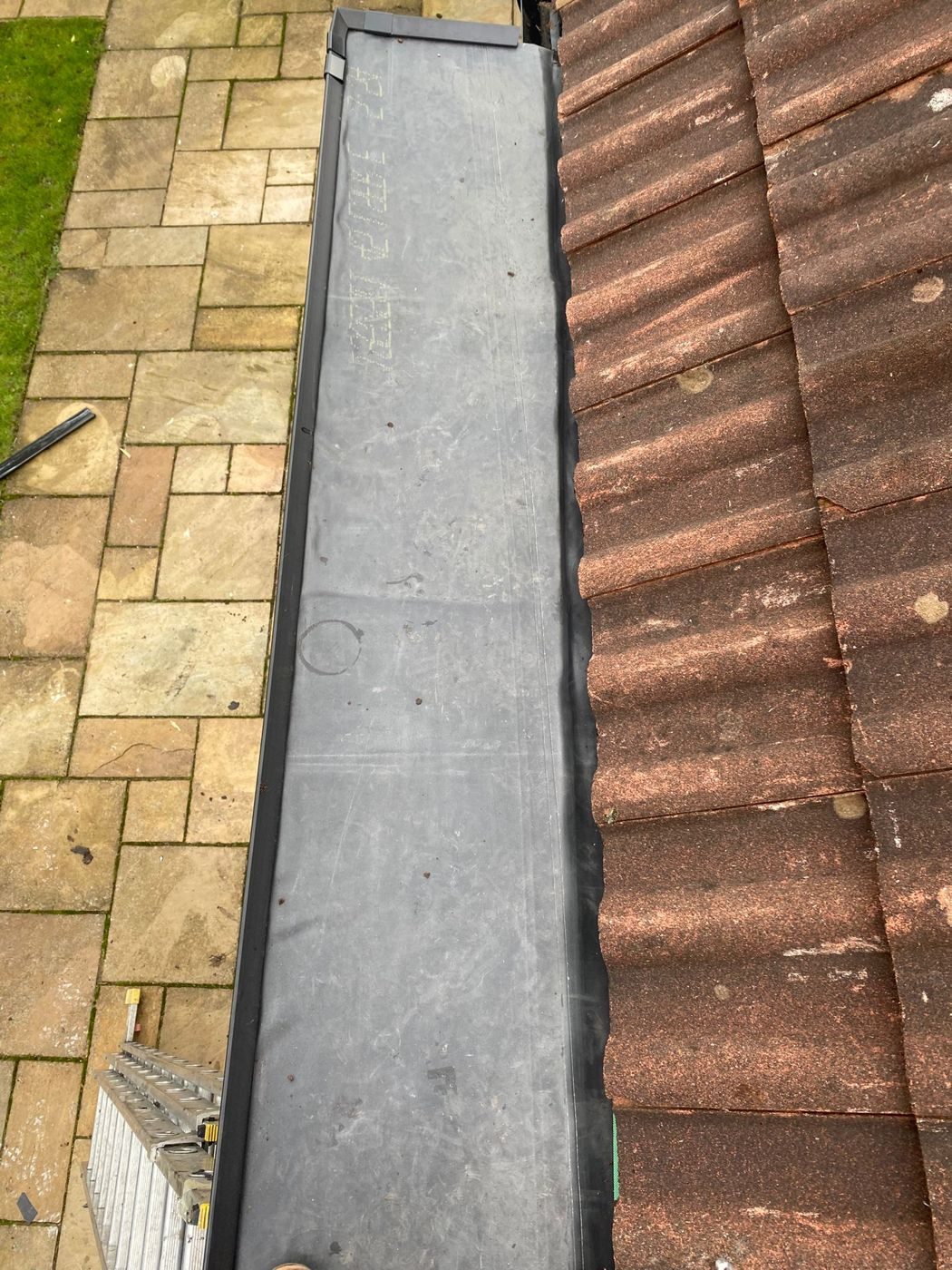 A finished sheet covering to a narrow lean-to roof alongside brown pantiles, with the black trim run along the edge
