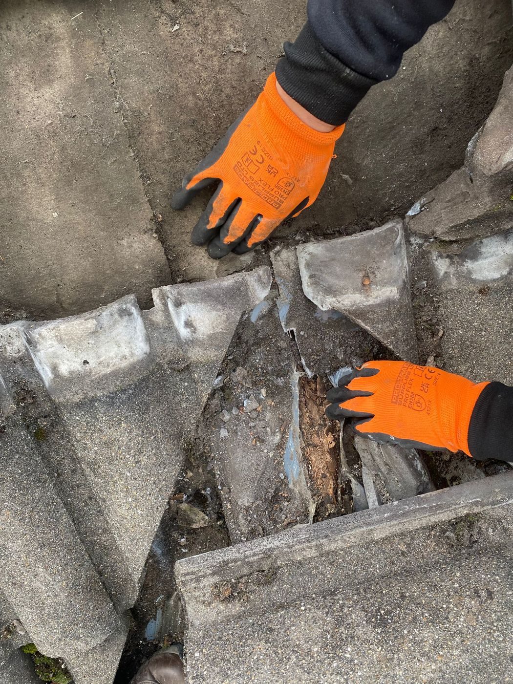Gloved hands lifting the tiles beside a failed valley, revealing rotten timber, split lining and packed debris underneath